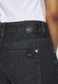 I jeans in denim scuro presentano un'alta vita, una toppa in pelle nera con il nome del marchio e cuciture doppie sulle tasche posteriori. Inclusi dettagli in metallo.