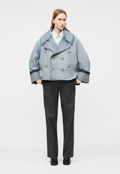 Frau steht mit den Händen in den Taschen, trägt eine hellblaue übergroße Jeansjacke, ein weißes Hemd mit Kragen, dunkle Hosen und schwarze Schuhe.