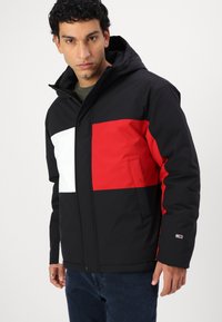 Veste à capuche noire et rouge avec des accents blancs, dotée d'une fermeture éclair à l'avant, de deux poches latérales et d'un logo sur la manche. Tissu lisse, résistant à l'eau.
