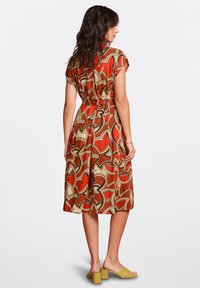 Short-sleeve, A-line dress in rust with abstract cream and brown pattern. Features a cinched waist and falls to mid-calf. Worn with green mules.