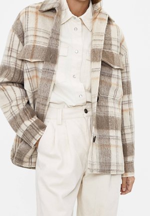 Kvinde iført beige og brun ternet jakke over cremefarvet skjorte med knapper og cremefarvede bukser med høj talje, med den ene hånd i lommen.