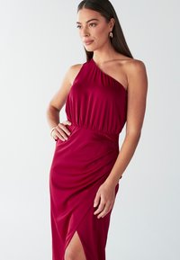 Vīnsarkan sarkana viendaļīga kleita no gluda auduma, ar savāktu vidukli un sānu spraugu. Izcelta ar smalkām drapējuma detaļām.