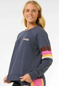 Marineblauwe sweatshirt met raglanmouwen, voorzien van kleurrijke gestreepte accenten in roze, bruin en geel, en een logo op de borst.