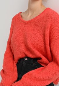 Maglione lavorato a maglia color corallo con vestibilità ampia e scollo ampio, caratterizzato da una trama a coste e maniche lunghe, abbinato a jeans neri a vita alta.