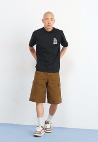 Sort kortærmet t-shirt med grøn lomme-detalje, parret med khaki cargo shorts og brune sneakers, præsenteret mod en neutral baggrund.