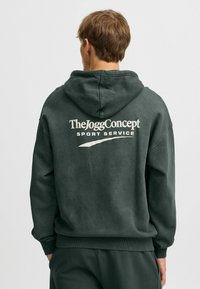 Felpa con cappuccio verde scuro dal tocco morbido. Presenta un logo sul retro "TheJoggConcept SPORT SERVICE" in bianco e un cappuccio regolabile con cordino.