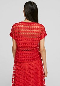 Top rojo transparente con recortes geométricos, mangas cortas y un escote redondeado, acompañado de una falda a juego con un diseño similar.