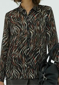 Blusa a maniche lunghe in tessuto trasparente con una base nera e motivi ondulati astratti bianchi e rossi. Ha una chiusura con bottoni e colletto.