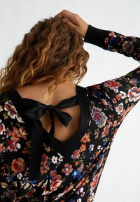 Černý květinový svetr s dlouhými rukávy, s výstřihem do V a stahovací stuhou na zádech. Zobrazuje barevné květinové vzory na texturované tkanině.