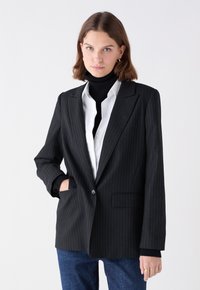 Blazer negro de rayas con solapa muesca, cierre de un botón y bolsillos frontales. Llevado sobre un cuello de tortuga negro y una camisa blanca.