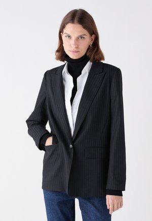 Blazer nero con righe verticali, colletto a revers, chiusura a un bottone e tasche frontali. Indossato su un dolcevita nero e una camicia bianca.