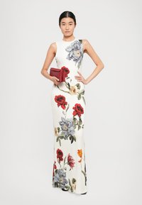 Woman in sleeveless white floor-length dress with large red, blue, and beige floral prints, holding a small red clutch bag.