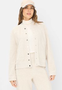 Beige Jacke mit Druckknopfverschluss aus strukturiertem Stoff, mit zwei aufgesetzten Taschen und einer lockeren Passform, kombiniert mit einem passenden Oberteil und einer Hose.