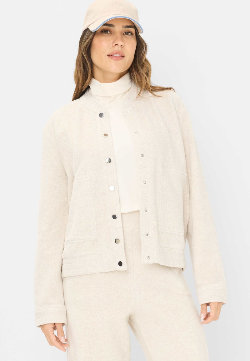 Beige Jacke mit Druckknopfverschluss aus strukturiertem Stoff, mit zwei aufgesetzten Taschen und einer lockeren Passform, kombiniert mit einem passenden Oberteil und einer Hose.