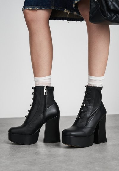 Juodos odinės platforminės aulinės botai su masyviu kulnu. Yra užtrauktukas ir dekoratyvi raištelių uždėjimo priekinė dalis su metaliniais akcentais. Tekstūruotas paviršius.