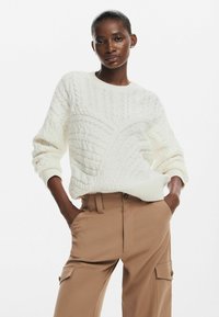 Maglione cream a lavorazione a trecce con un motivo testurizzato, colletto rotondo e polsini a costine, abbinato a pantaloni khaki con tasche cargo.