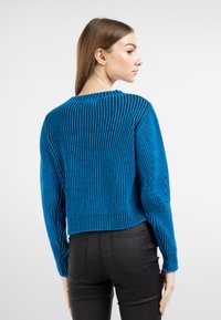 Maglione blu corto con texture a coste verticali e maniche lunghe, dotato di un collo rotondo e dettagli a maglia leggermente aperti. Abbinato a jeans neri.