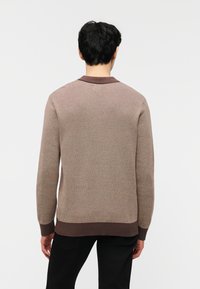 Άνδρας με μαύρα μαλλιά που φοράει ένα μακρυμάνικο καφέ πλεκτό πουλόβερ με σκουρό καφέ γιακά και τελείωμα, γυρισμένος προς τα πίσω μπροστά σε απλό φόντο.