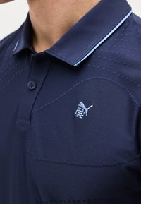 Κοντινό πλάνο για το γιακά μπλε ναυτικού polo με γαλάζια επένδυση και μικρό κεντημένο λογότυπο Puma στο στήθος.