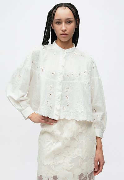 Fehér, rövidített blúz virágos hímzéssel, gombos záródással és fodros elülső résszel. Hosszú ujjú, laza fazonú, hullámos szegélyű.