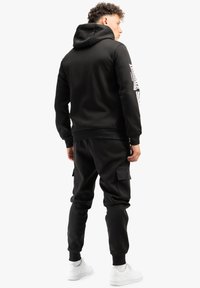 Melnā kapučas džemperis ar brīvu piegriezumu, kas rotāts ar logotipu uz labā piedurknes, apvienots ar melniem sporta biksēm ar kabatām.