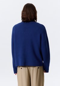 Pull en bleu marine avec une coupe ample, un col côtelé et des manches longues, associé à un pantalon beige clair. Texture douce, détails minimaux.