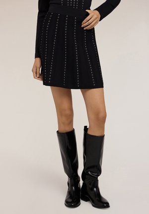 Mini gonna nera con fila verticale di borchie metalliche, abbinata a stivali neri lucidi alti. Tessuto a maglia testurizzato, design aderente.