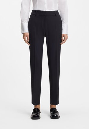 Donna che indossa pantaloni formali neri a gamba dritta, camicia bianca a manica lunga con bottoni e mocassini neri, in piedi su sfondo bianco.