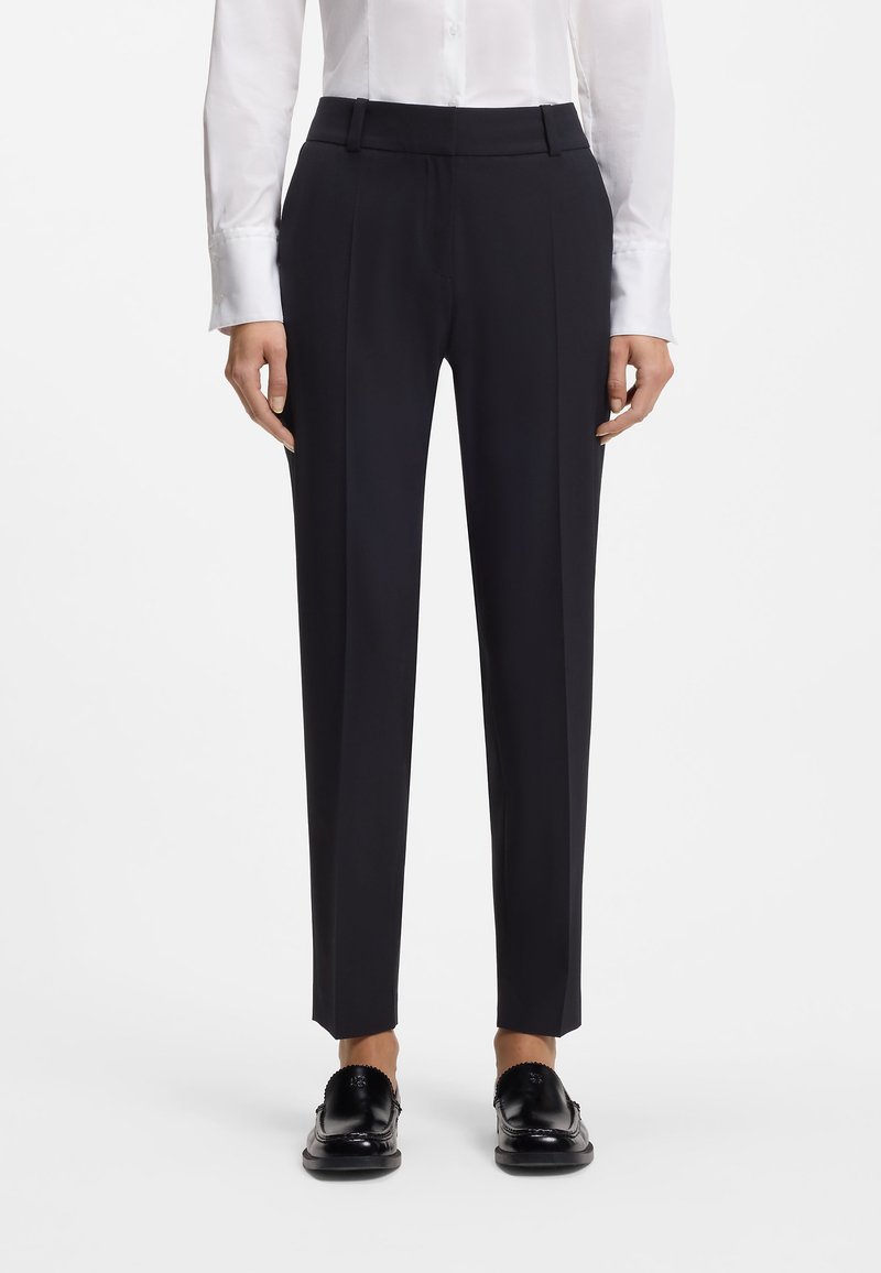 Femme portant un pantalon formel noir à jambes droites, une chemise blanche à manches longues boutonnée et des mocassins noirs, debout devant un fond blanc.