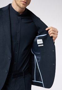 Uomo in abito slim fit blu scuro e camicia con colletto a bandiera tiene aperto il giubbotto per mostrare l'inserto in raso blu e le etichette del marchio.