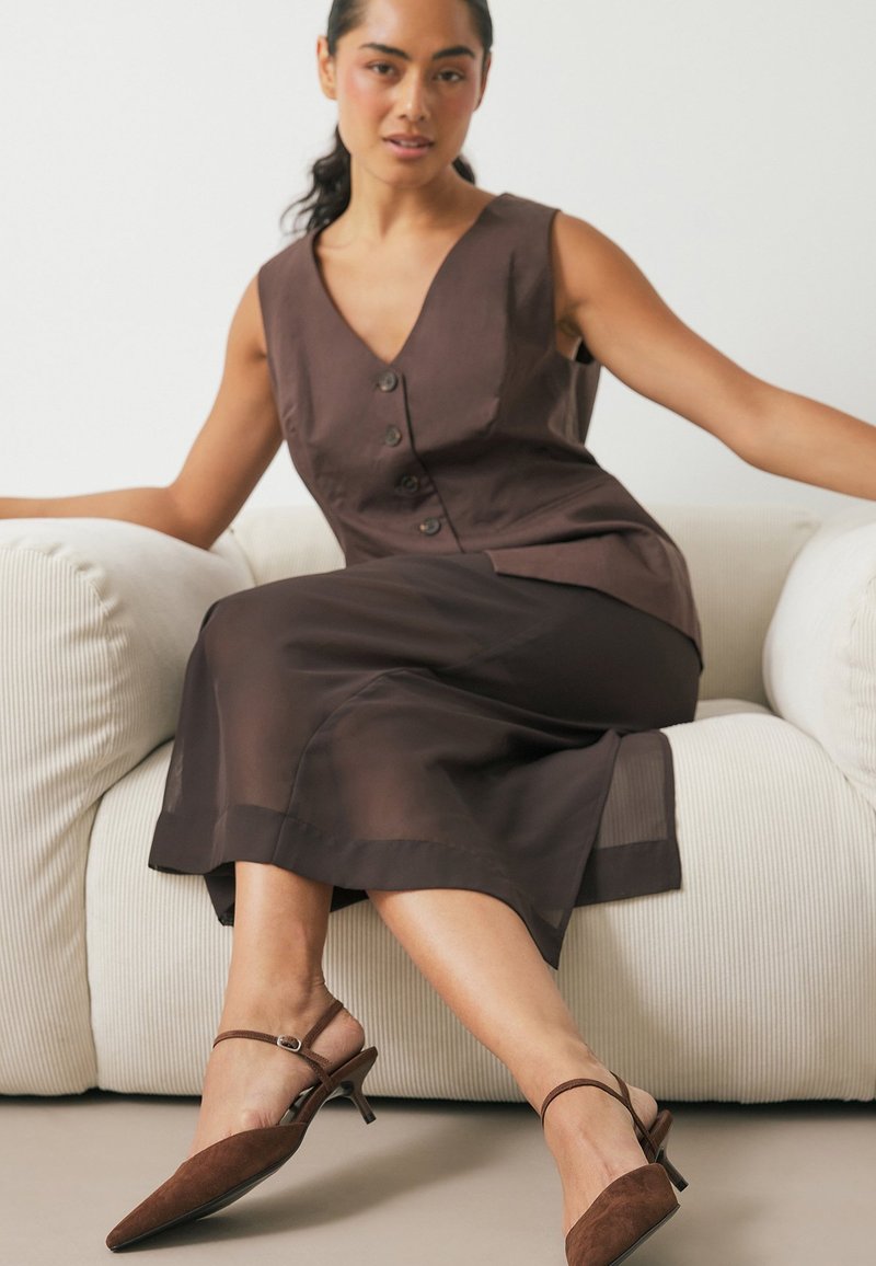 Rjava brezrokavna obleka z detajli gumbov, zložena preko prosojne rjave krila. Rjavi semiš slingback petki s konicami.