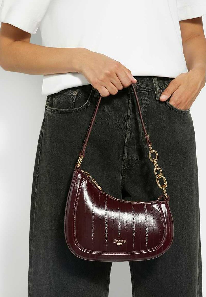Burgundy patent leather handbag with a curved shape, gold chain accent, and zip closure, held by a person wearing a white shirt and black pants.