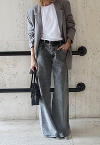 Personne portant un blazer gris, un t-shirt blanc, un jean gris à jambes larges, tenant un sac à main noir, debout devant un mur beige carrelé avec des rampes noires.