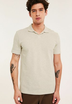 Junger Mann mit lockigem Haar, der ein beiges kurzärmeliges Poloshirt trägt und Tätowierungen eines Vogels und eines Schmetterlings auf seinen Unterarmen zeigt.