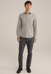 Chemise à manches longues gris clair avec un col boutonné, associée à un pantalon écossais sombre et des chaussures noires à lacets. Texture lisse, coupe ajustée.