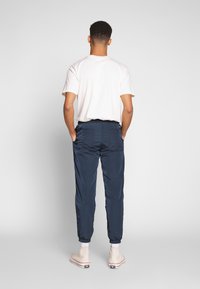 Man med vit t-shirt, marinblå joggers med elastiska muddar, vita strumpor och off-white högklackade sneakers, stående med händerna i fickorna.