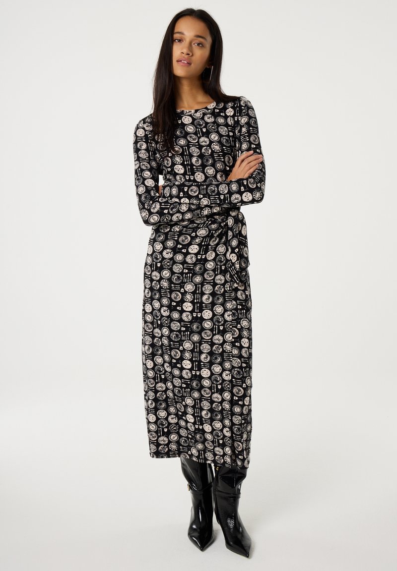 Schwarzes Langarmkleid mit einem kreisförmigen Muster in Weiß. Figurbetonte Silhouette, knielang, kombiniert mit glänzenden schwarzen kniehohen Stiefeln.