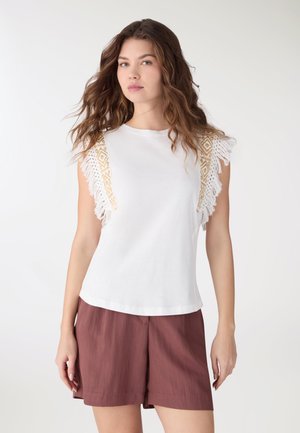 Giovane donna con capelli sciolti e ondulati, indossa un top bianco senza maniche con frange e pantaloncini marroni a vita alta su sfondo bianco.