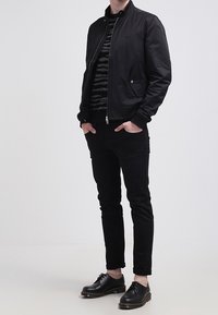 Czarna kurtka bombowca z zapięciem na zamek, zestawiona z pasiastym swetrem w czarno-szarych odcieniach, czarnymi spodniami typu slim-fit oraz czarnymi butami na sznurowadła.