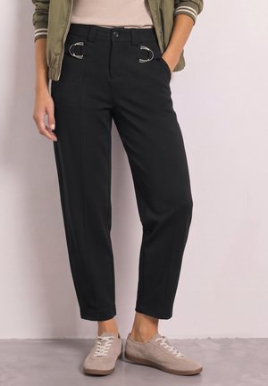 Personne portant un pantalon court noir avec des détails de boucle métallique, des baskets beiges, un haut blanc et une veste vert olive, debout contre un mur uni.