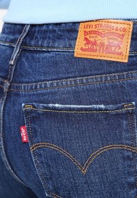 Jeans de mezclilla azul oscuro con un parche de cuero marrón con el logo de Levi Strauss, una etiqueta roja en el bolsillo trasero y costuras amarillas en contraste.