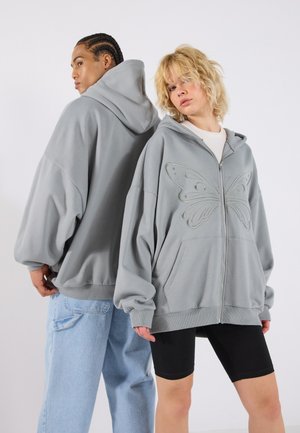 Γκρίζες ζακέτες με φερμουάρ και oversized εφαρμογή, με κεντημένο μια πεταλούδα στο μπροστινό μέρος. Δύο τσέπες, ριμπ μανσέτες και απαλή υφή.