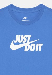 Mėlyna medvilninė marškinėliai su balta „Just Do It“ fraze, į kurią įkomponuota Nike Swoosh simbolika. Klasikinio apvalaus kaklo dizainas.