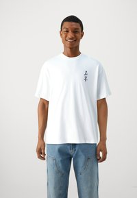 White cotton t-shirt with short sleeves, featuring a black graphic design on the left chest. Paired with light blue loose-fitting jeans.
