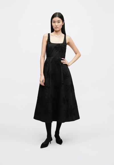 Μαύρο άνετο φόρεμα χωρίς μανίκια με κεντημένα λουλουδάτα σχέδια, εφαρμοστό κορσάζ και ρέουσα φούστα. Συνοδεύεται από μαύρες καλσόν και尖 πόδια παπούτσια.