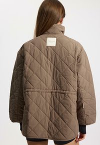 Brown quilted jacket with a high collar, featuring a square label at the back, and a loose fit. Smooth fabric with diagonal stitching patterns.