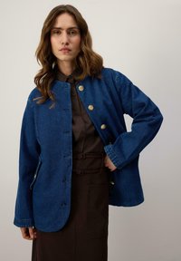 Modrá džínová bunda s okrúhlym golierom, zlatými gombíkmi a bočnými vreckami. Má dlhé rukávy a voľný strih. Nosená cez tmavohnedý top.