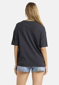 Γυναίκα με μακριά ξανθά μαλλιά που φοράει ένα χαλαρό μαυρο κοντομάνικο μπλουζάκι και ανοιχτό μπλε τζιν σορτσάκι, γυρισμένη προς τα πίσω σε λευκό φόντο.