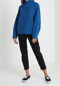 Pull à col roulé bleu tricoté avec un motif texturé, associé à un jean noir droit et des baskets noires, posé sur un fond clair.