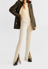 Olivengrøn parka med pelsforet hætte, beige top og cremede bukser med flared ben og splitkanter, parret med sorte ankelstøvler.